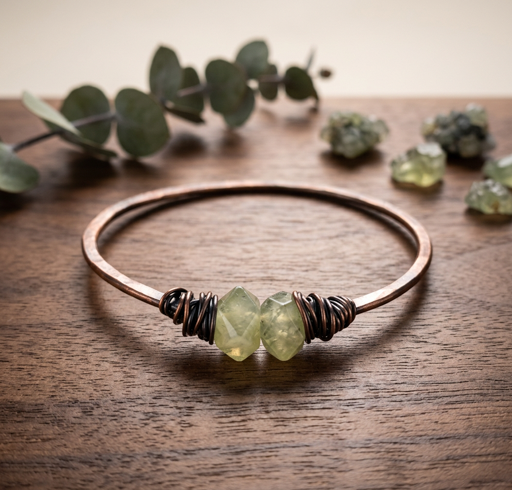 Warm metal-tone bangle with pale green irregular-cut crystalline stones and dark coiled accents on a wooden surface.