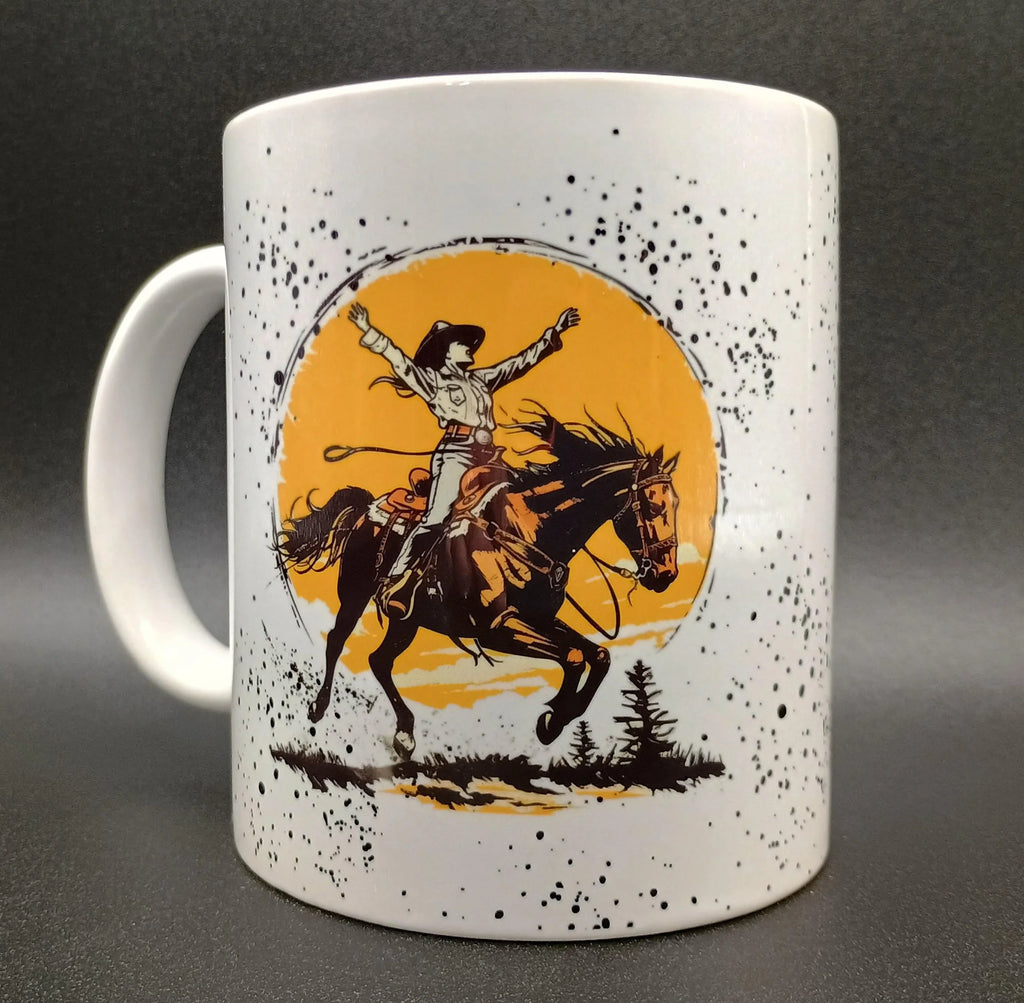 White ceramic mug with black speckles featuring a cowgirl on a bucking horse against an orange sun