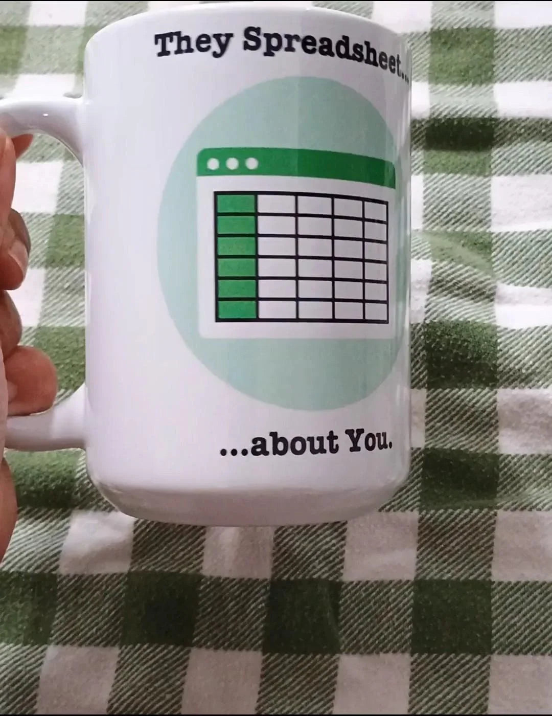 White coffee mug with spreadsheet graphic and text 'They Spreadsheet... about You' on green checkered tablecloth