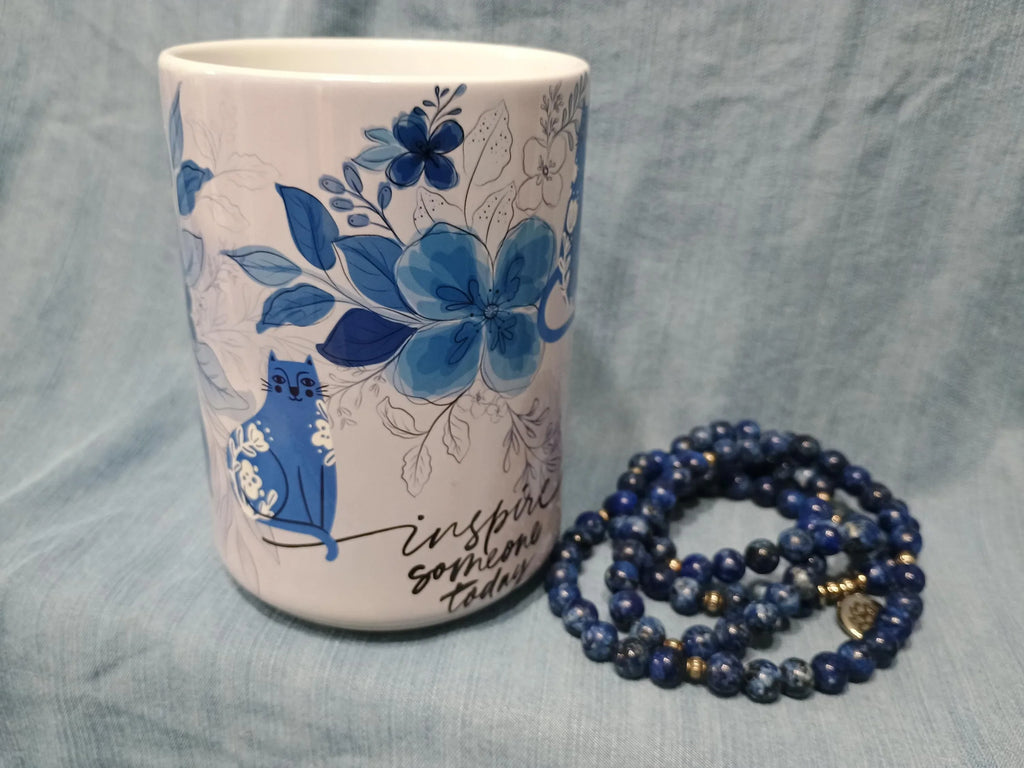 White ceramic mug with blue floral and cat design beside coiled blue beaded bracelet on light blue fabric