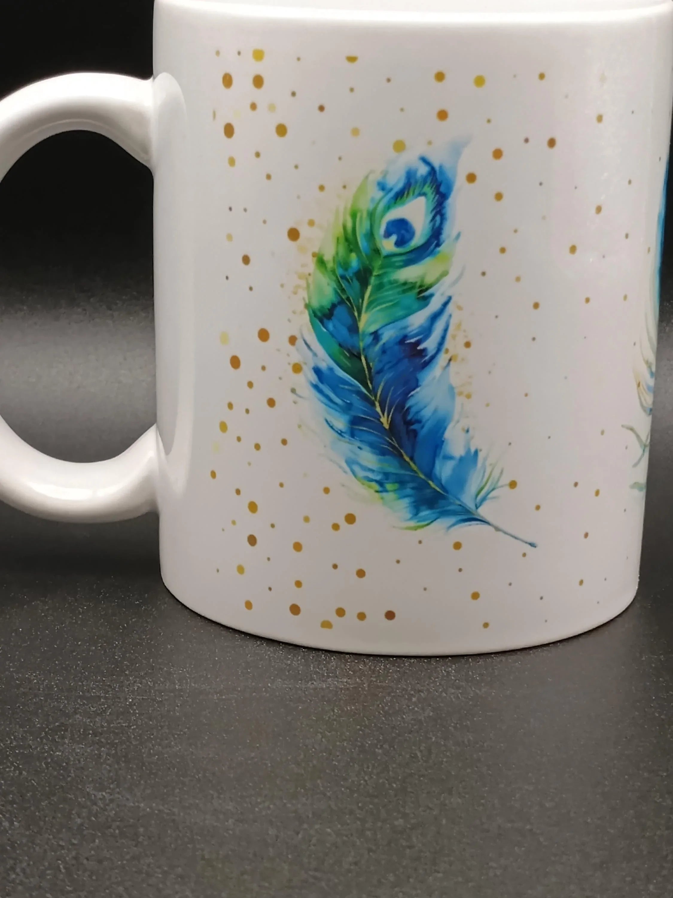 White ceramic mug with a colorful blue and green peacock feather design and gold dots
