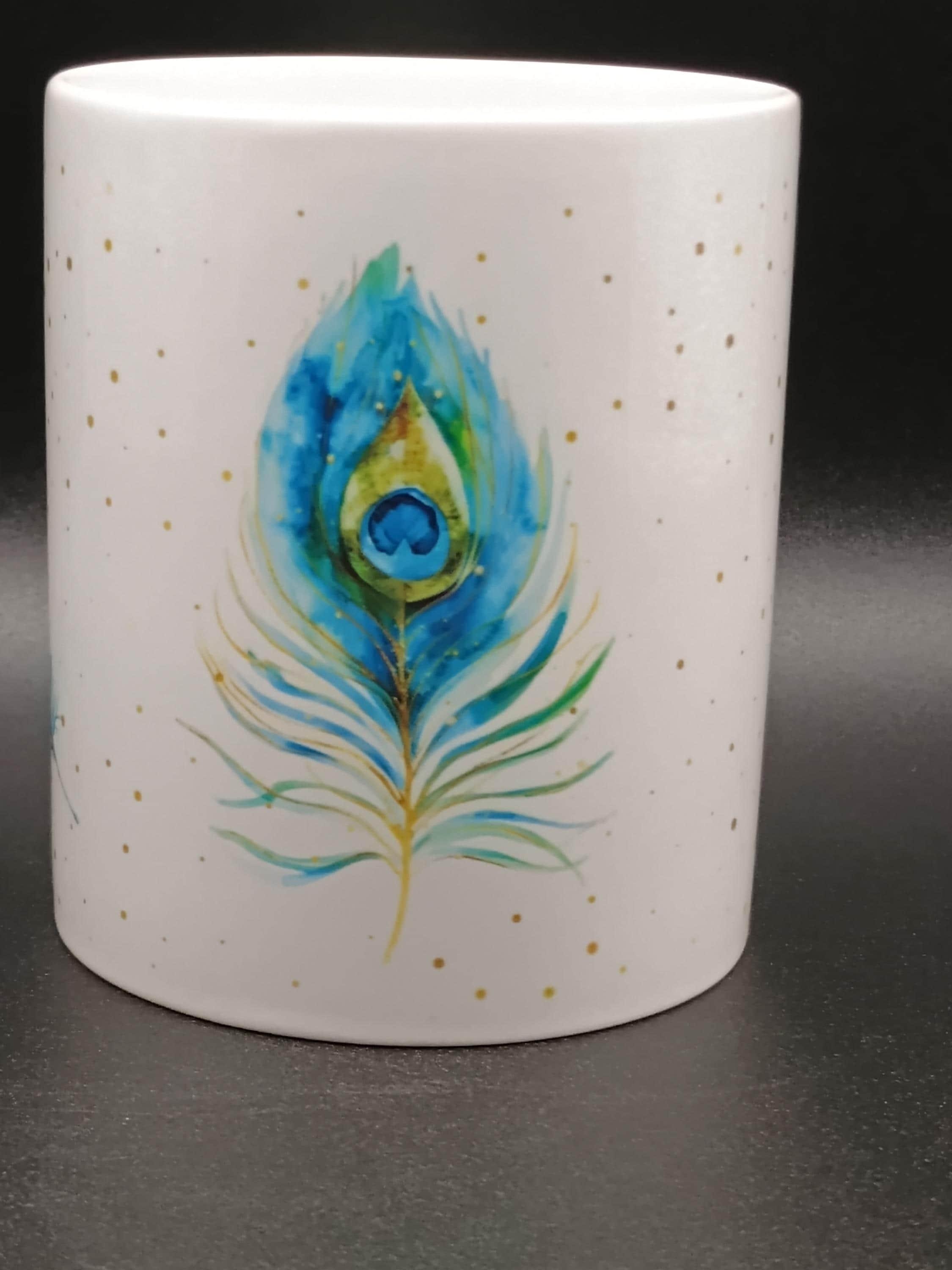 White ceramic mug with a blue and green watercolor peacock feather design and gold speckles