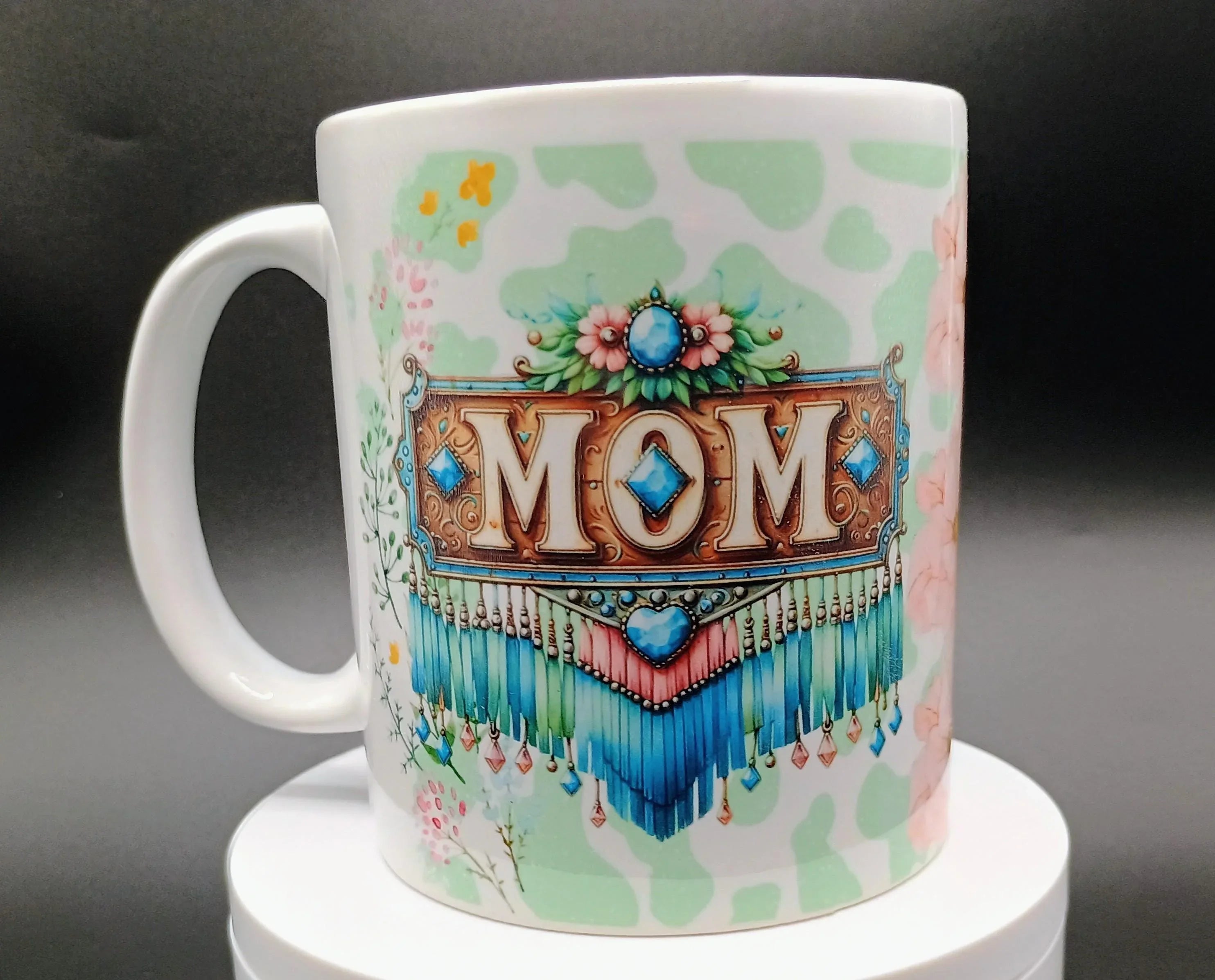 White ceramic mug with colorful floral and geometric design featuring the word MOM and decorative fringe