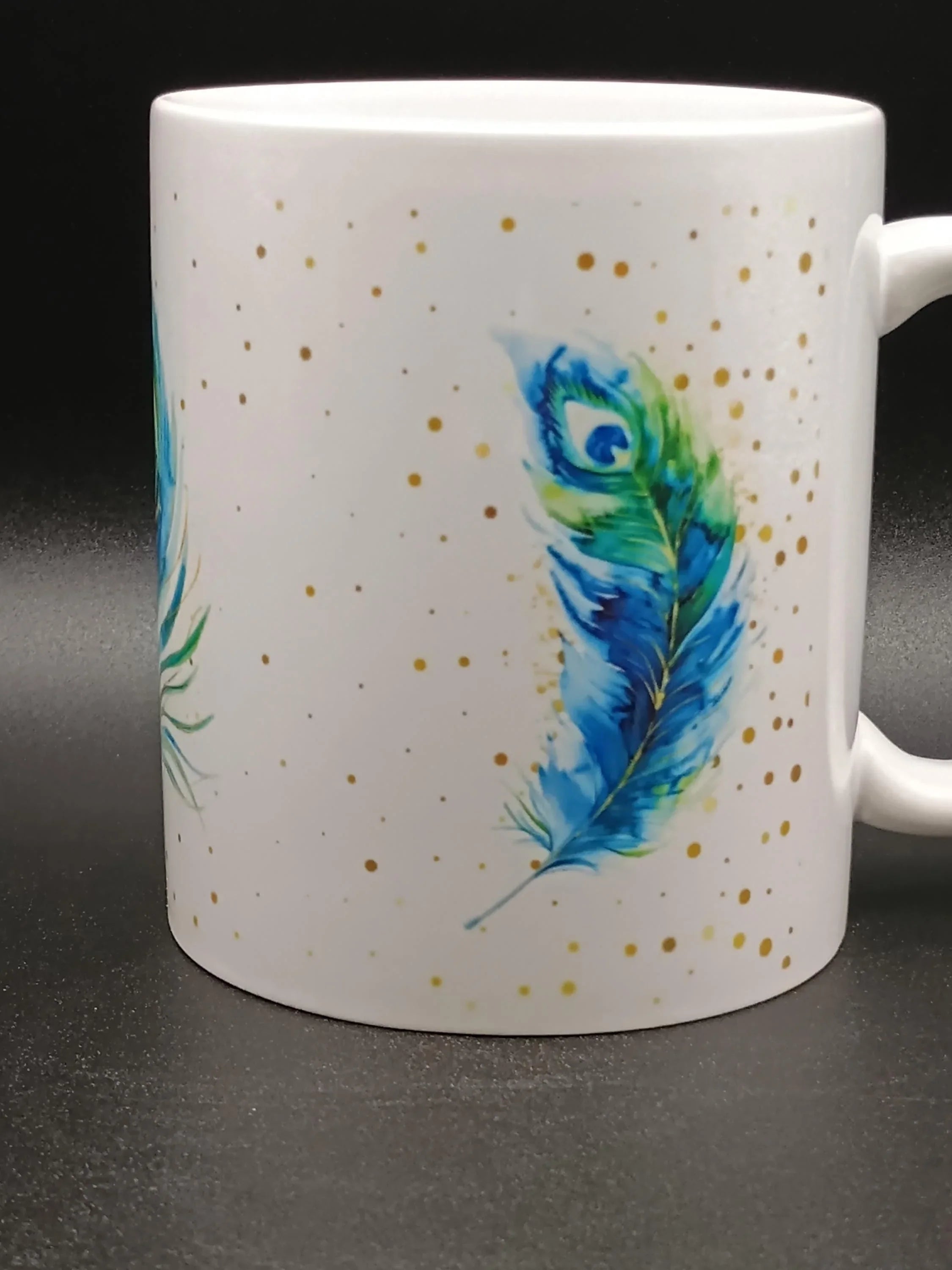 White ceramic mug with blue and green peacock feather design and gold speckles on black background