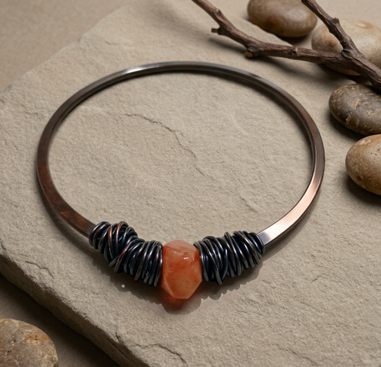 Oxidized copper bangle with an irregular-cut orange stone and dark coiled accents on a neutral stone surface.