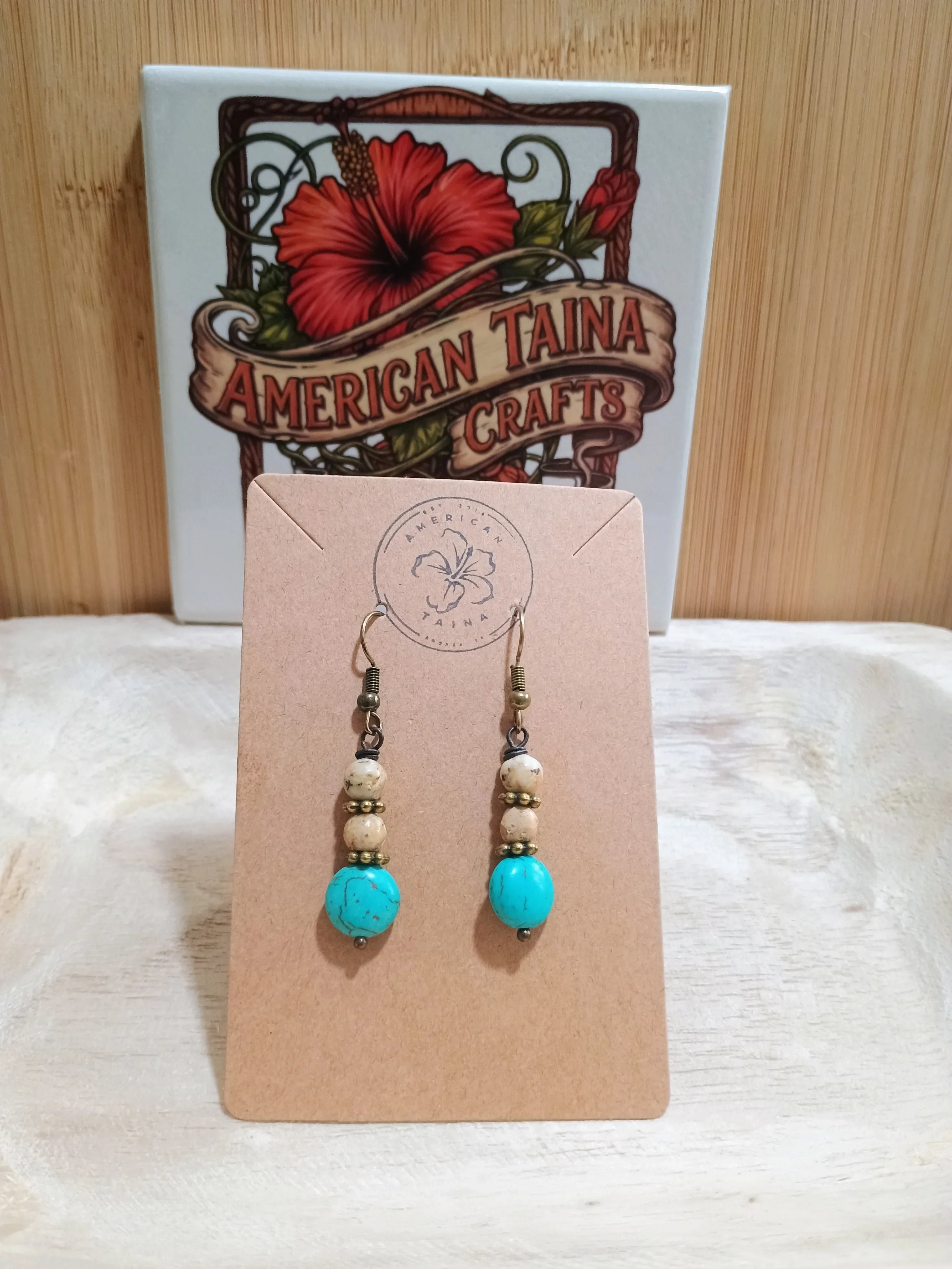 Handmade turquoise and beige beaded earrings on brown card with American Taina Crafts logo and wooden background