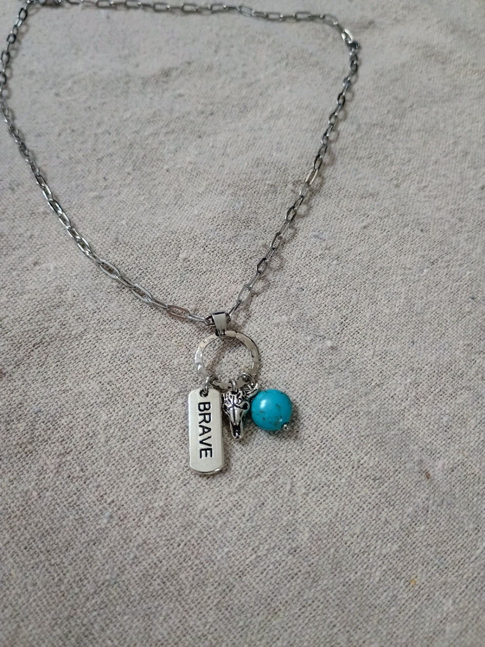 Silver chain necklace with circular pendant holding brave tag, bull head charm, and turquoise bead on beige fabric