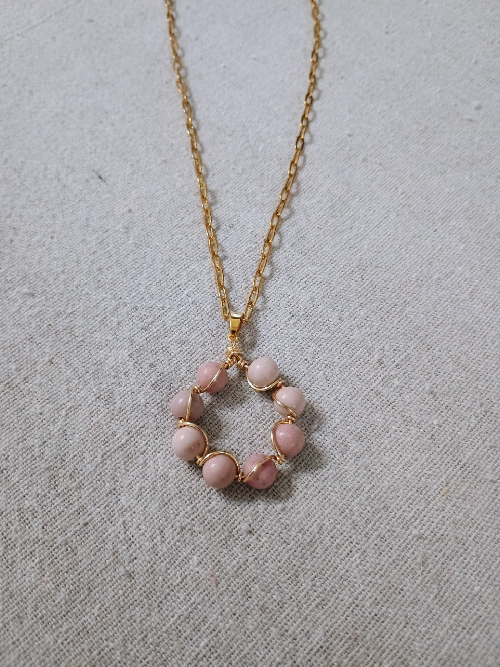 Handcrafted gold chain necklace with circular pendant made of pink beads wrapped in gold wire on textured fabric