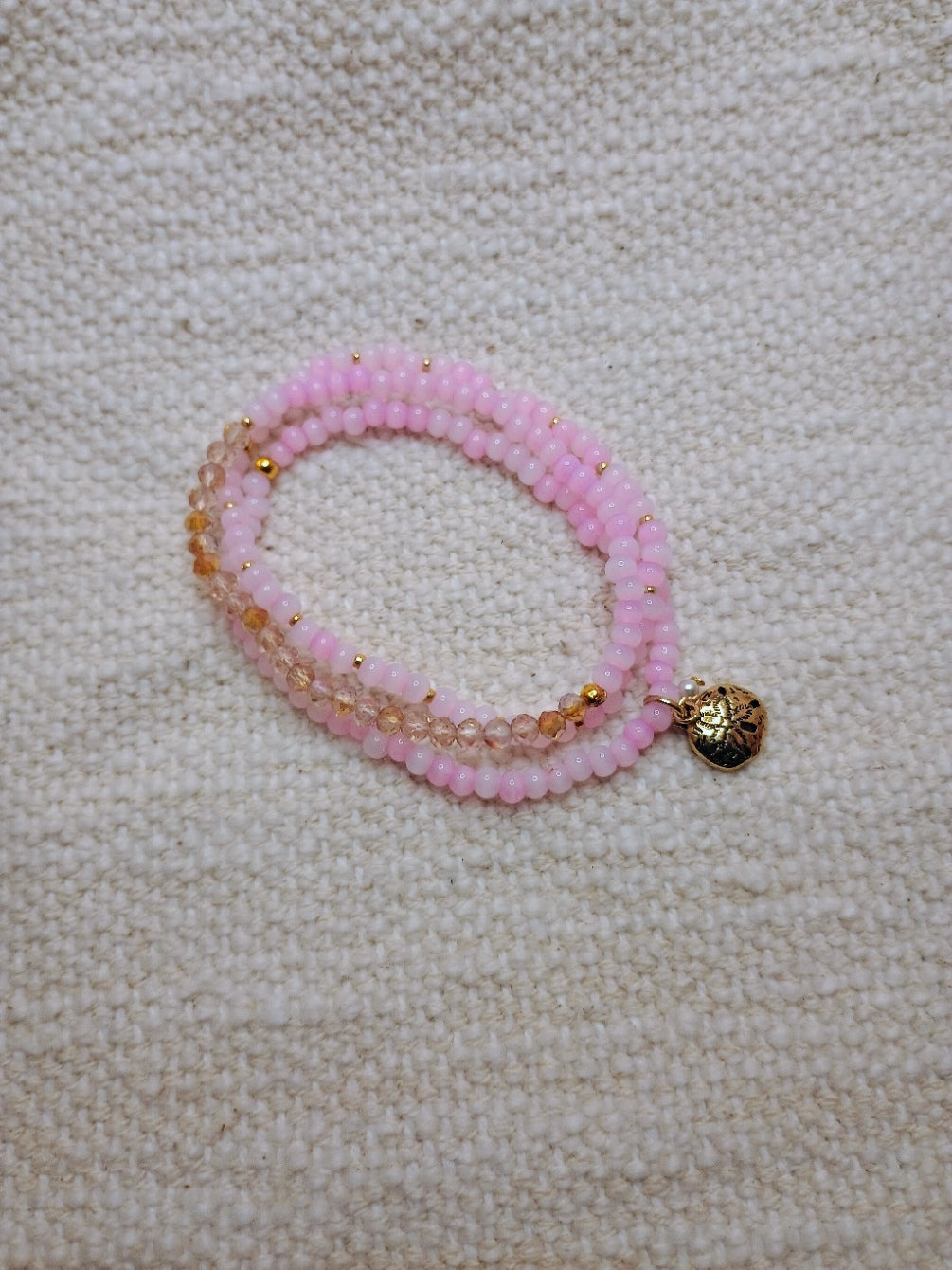 Pink beaded bracelet with a gold charm on a beige fabric background
