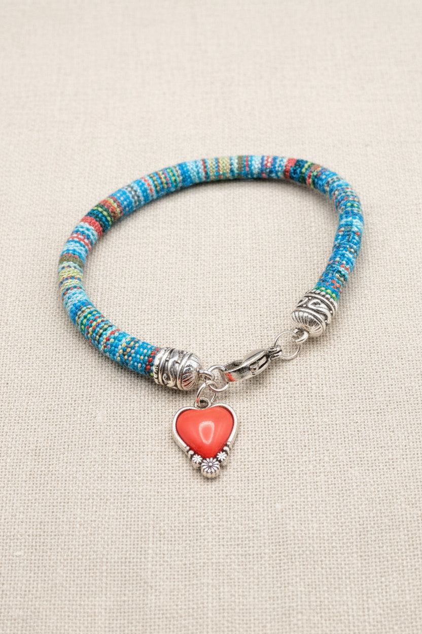 A beaded bracelet with a heart-shaped coral pendant, featuring a multicolor blue, red, and yellow beaded band, silver-toned metal spacers, and a silver heart charm on a dark gray background.