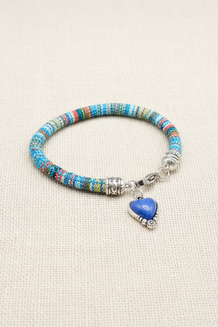 A beaded bracelet with a blue heart-shaped pendant, displayed on a dark gray background. The bracelet features a multicolor beaded pattern with turquoise, blue, red, and yellow beads, accented by silver-toned metal spacers and a dangling heart charm.