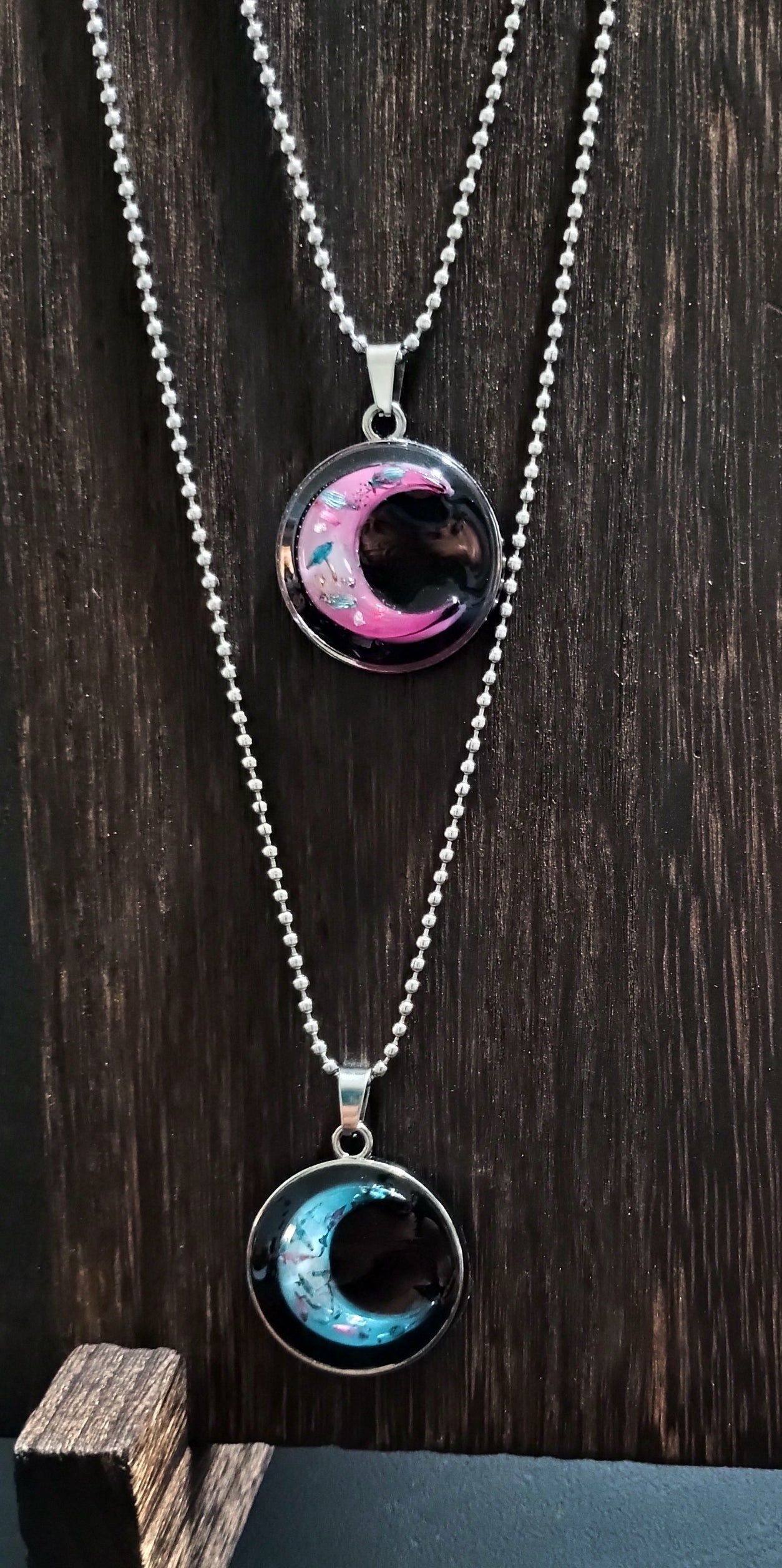 Two handmade crescent moon resin pendant necklaces on silver ball chains — one in cosmic pink and one in galaxy teal — displayed against a dark wood background