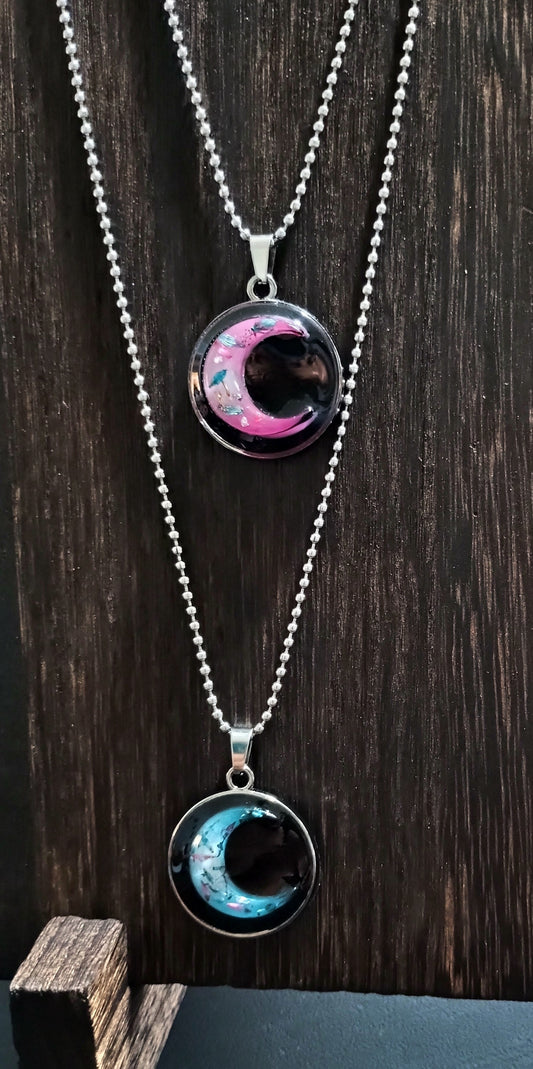 Two handmade crescent moon resin pendant necklaces on silver ball chains — one in cosmic pink and one in galaxy teal — displayed against a dark wood background