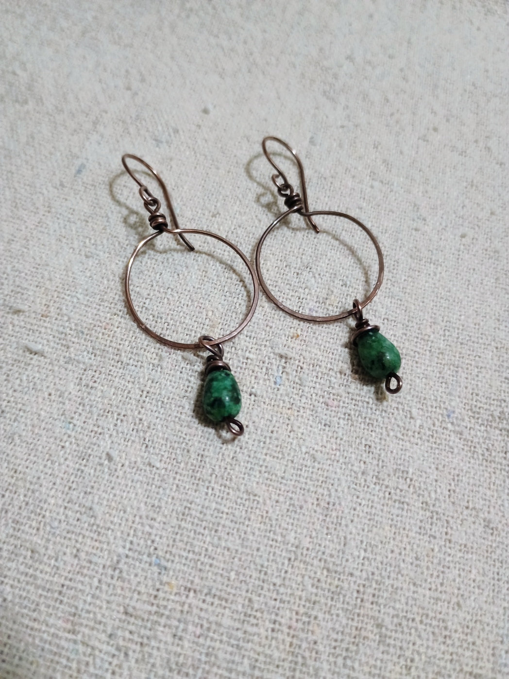 Oxidized copper hoop earrings with mottled green teardrop beads on light fabric.