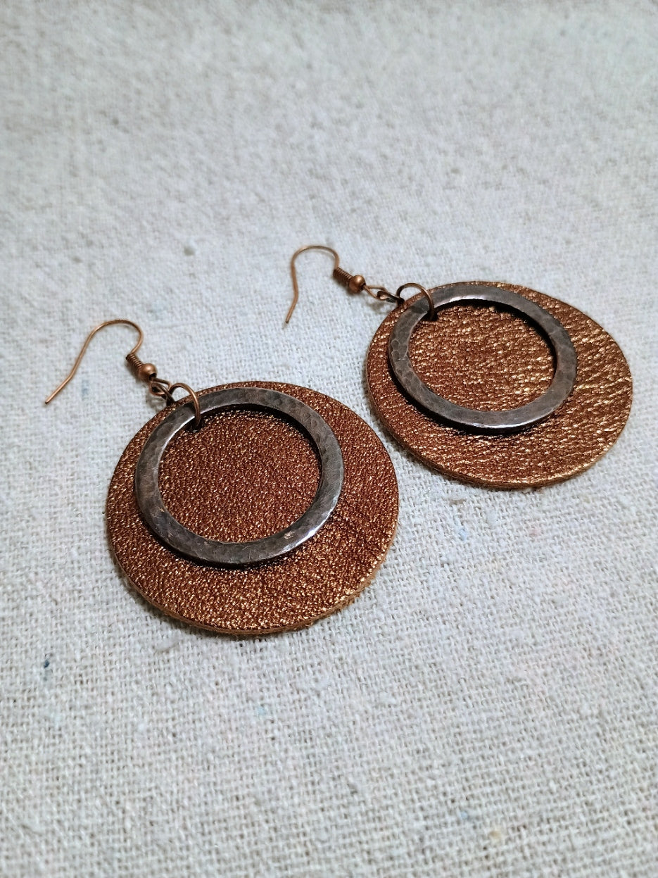Shimmering copper-colored leather circle earrings layered with oxidized copper rings on light fabric.