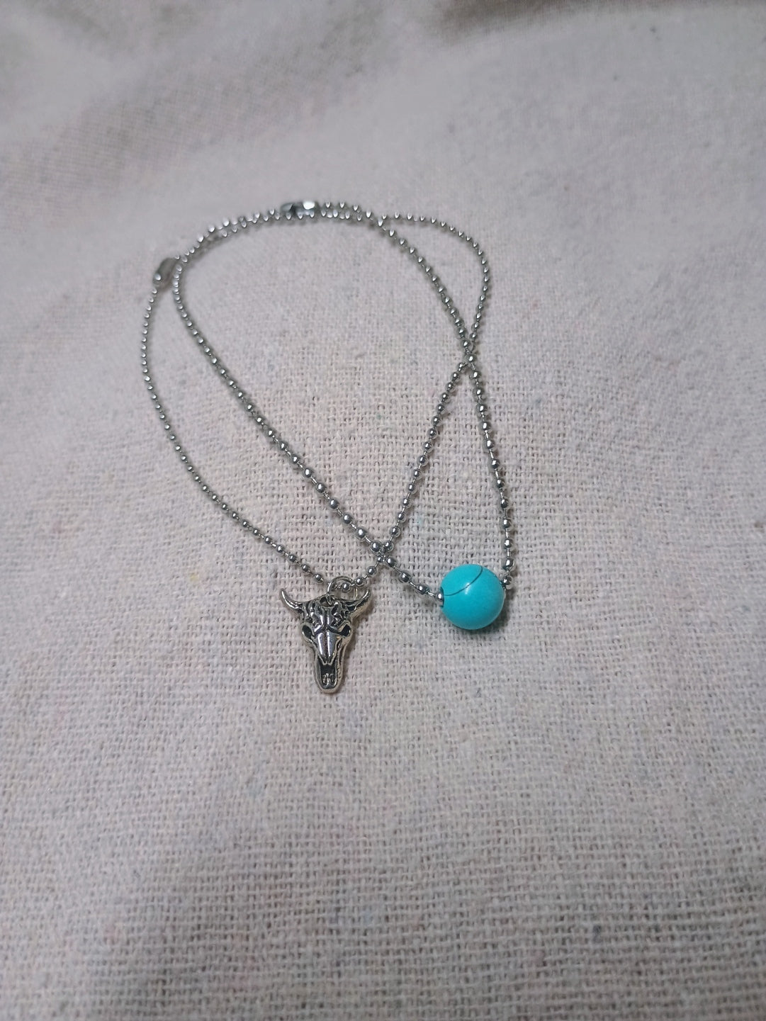 Two silver-tone ball-chain bracelets, one with a turquoise-colored bead and one with a bull-skull charm