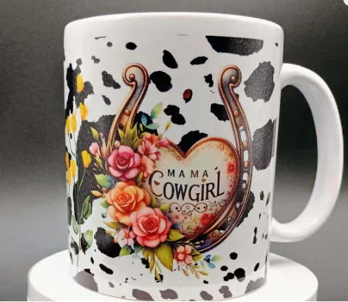 White ceramic mug with black cow spots, floral bouquet, horseshoe, and text Mama Cowgirl