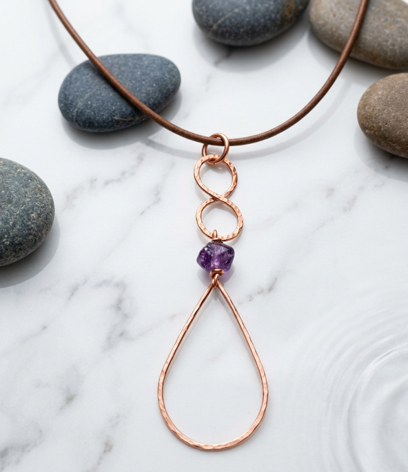 A copper wire pendant necklace with a teardrop-shaped loop, a small infinity symbol, a textured purple gemstone, and a simple copper cord, styled on a white marble surface with smooth gray stones.