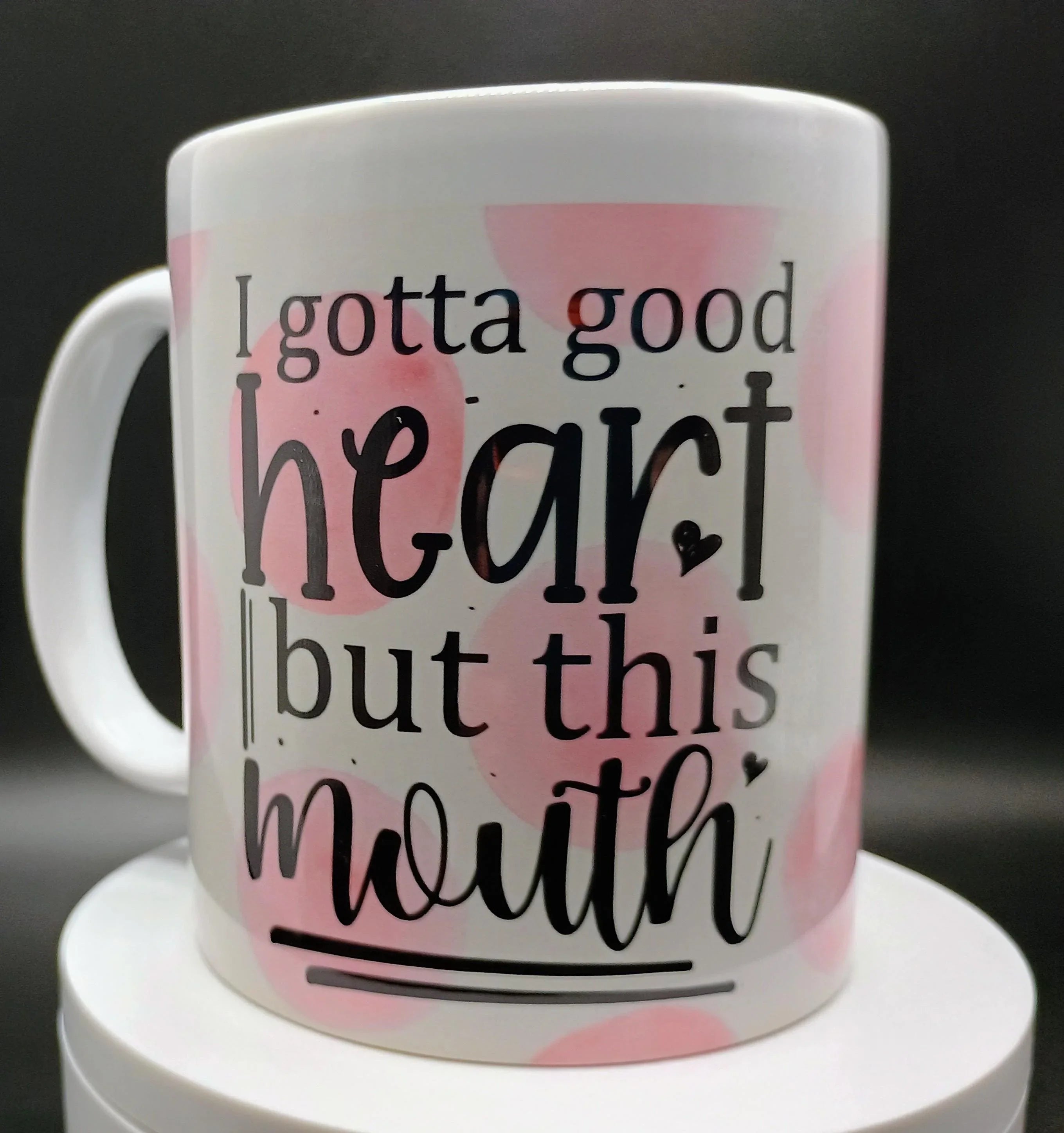 White ceramic mug with pink spots and black text reading 'I gotta good heart but this mouth'