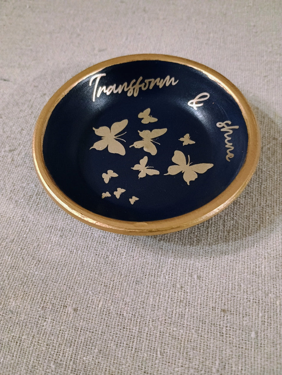 Handcrafted blue ceramic ring dish with gold butterflies and text 'Transform & shine' with gold rim on fabric surface