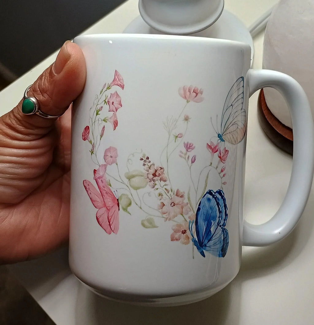 White ceramic mug with pink and blue butterfly and floral pattern held by hand with turquoise ring
