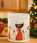 White ceramic mug with colorful cartoon cats wearing scarves, festive Christmas tree and bokeh lights background