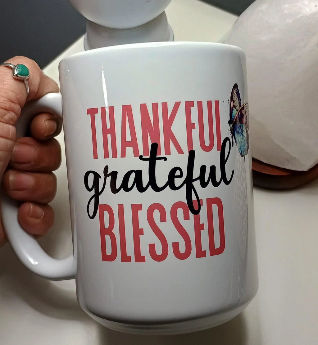 White ceramic mug with the words 'Thankful grateful blessed' in red and black, held by a hand with a turquoise ring