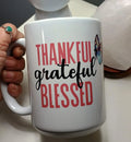White ceramic mug with the words 'Thankful grateful blessed' in red and black, held by a hand with a turquoise ring