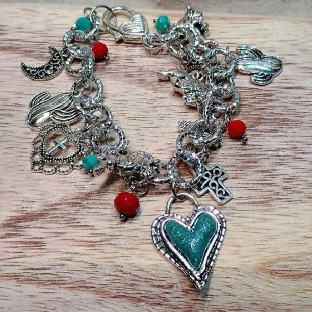 Silver charm bracelet with green heart pendant, turquoise and red beads, crescent moon and cross charms on wooden surface
