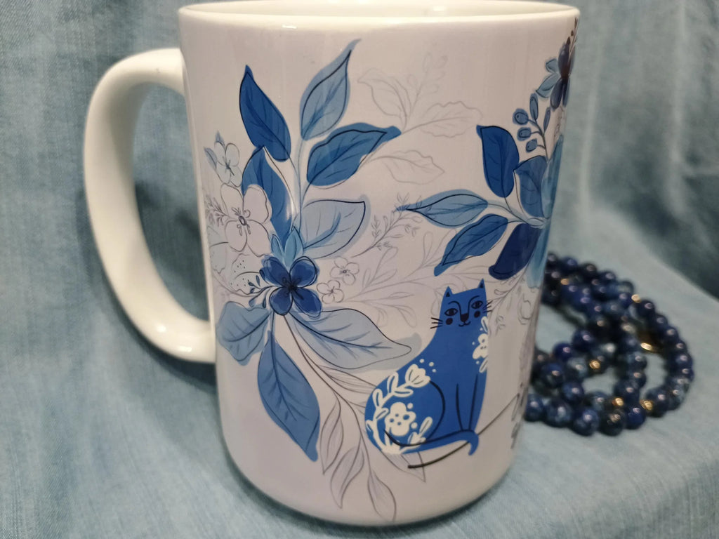 White ceramic mug with blue floral design and a blue cat illustration on fabric background