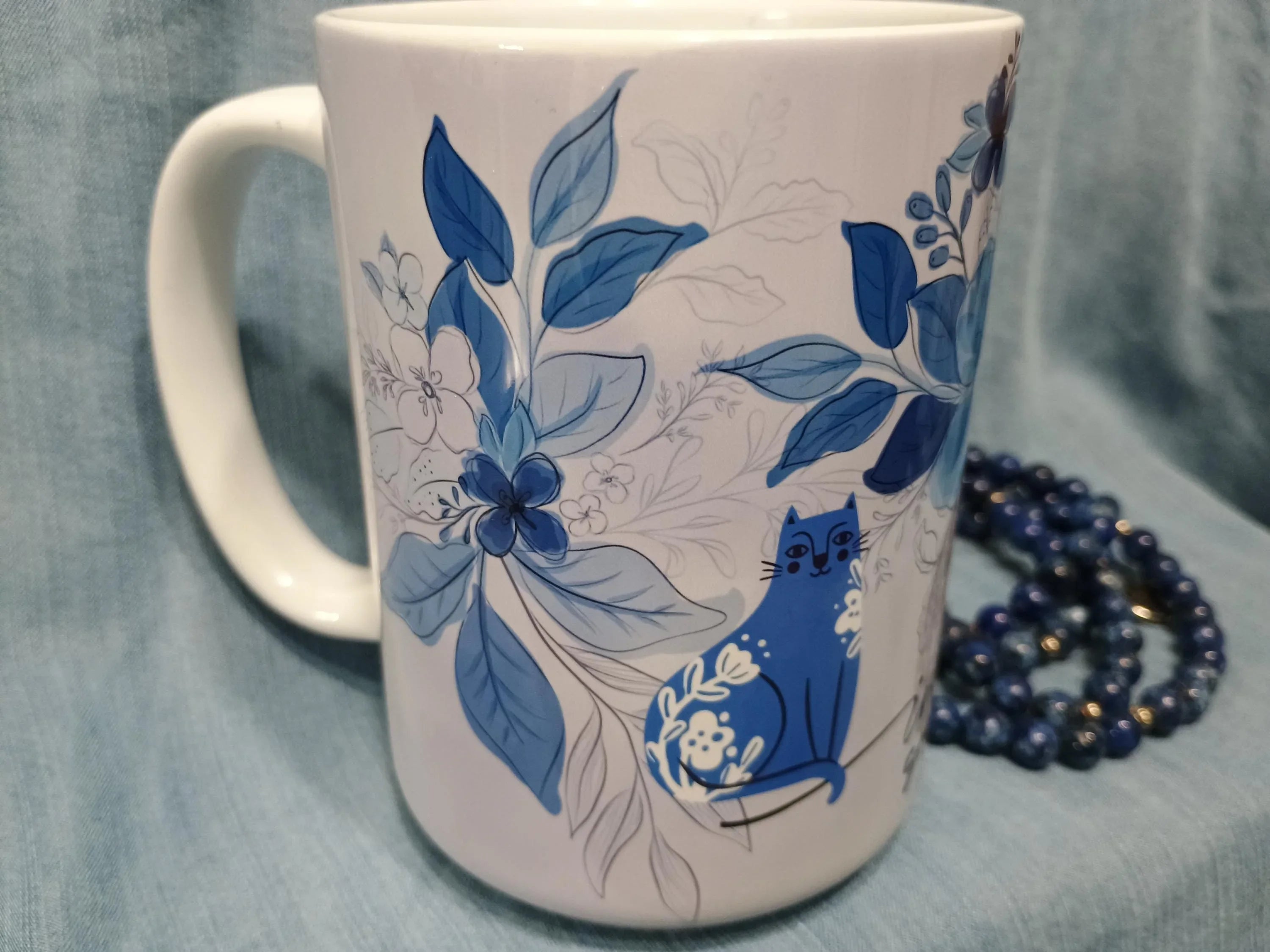 White ceramic mug with blue floral design and a blue cat illustration on fabric background