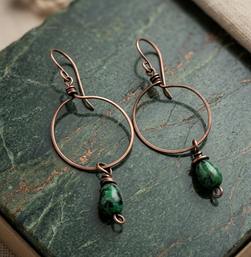 Copper hoop earrings with green teardrop beads on a dark green stone surface.