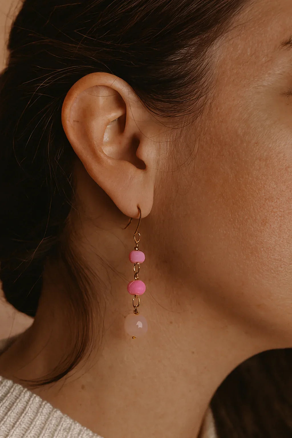 Close-up of woman's ear wearing gold dangle earrings with pink beads on neutral background