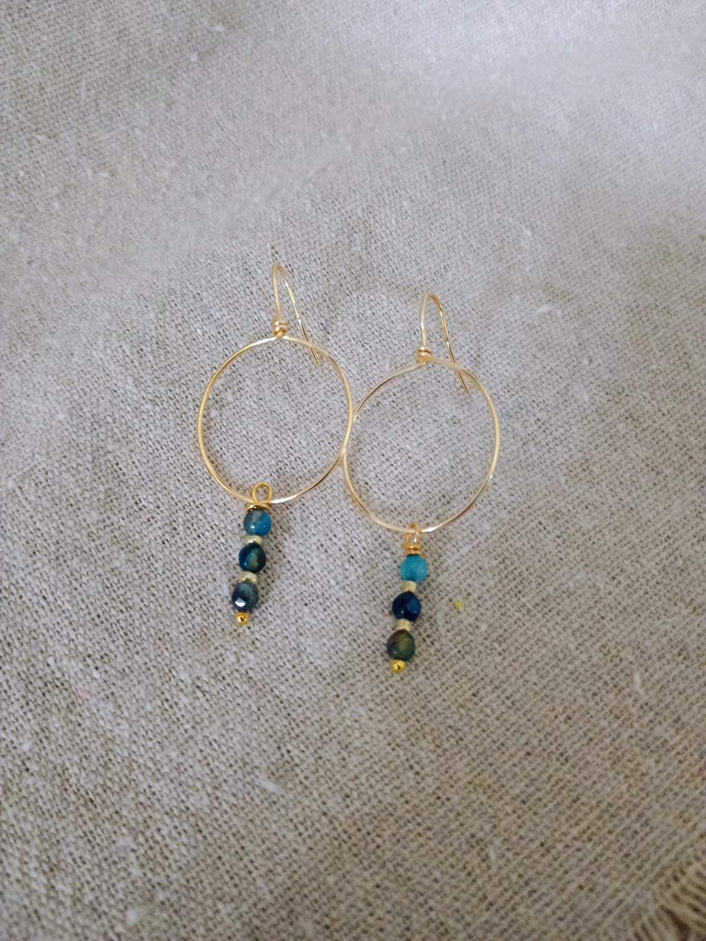 Gold hoop earrings with three small blue-green beads hanging, on beige fabric background