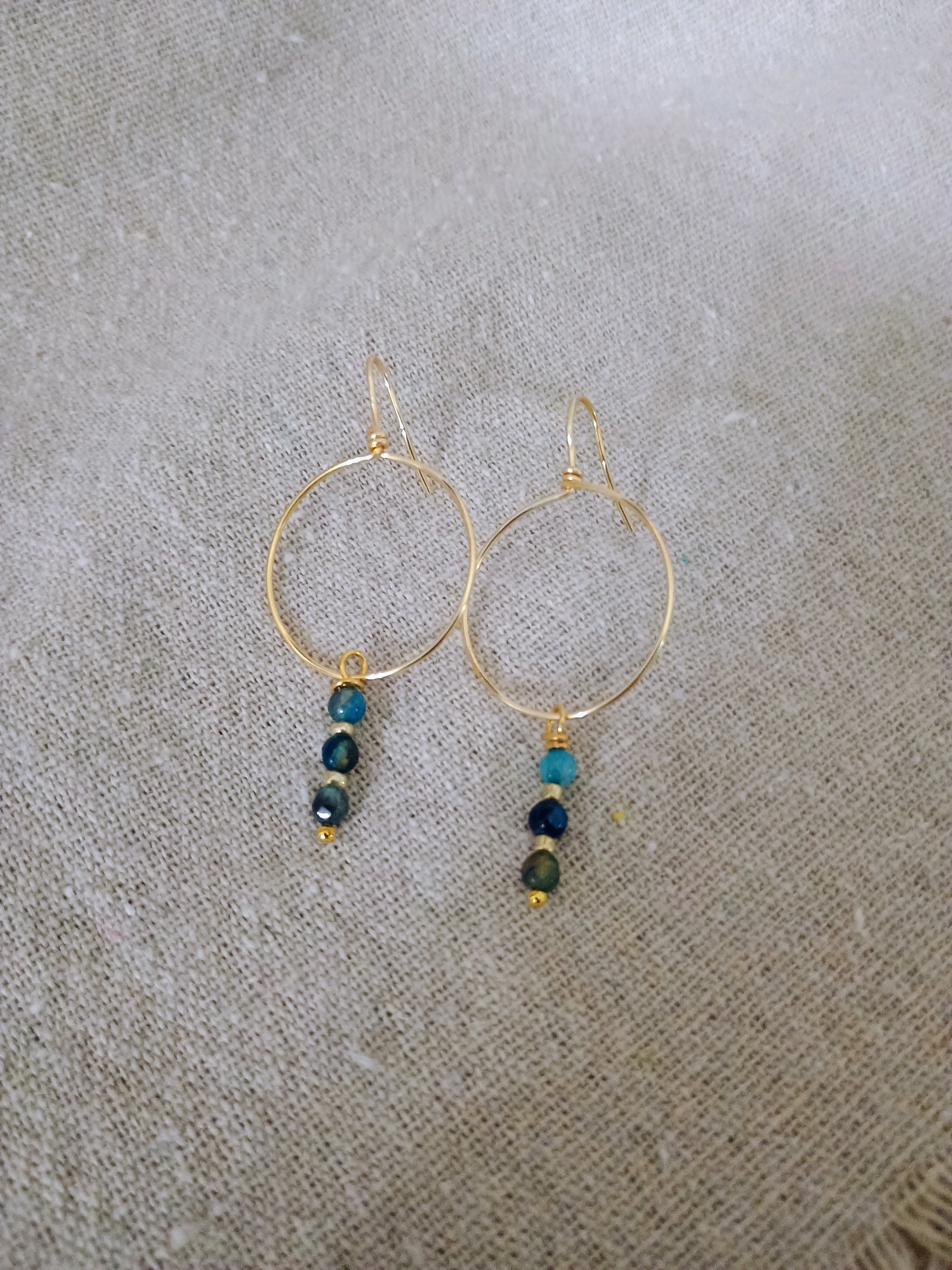 Gold hoop earrings with three small blue-green beads hanging, on beige fabric background