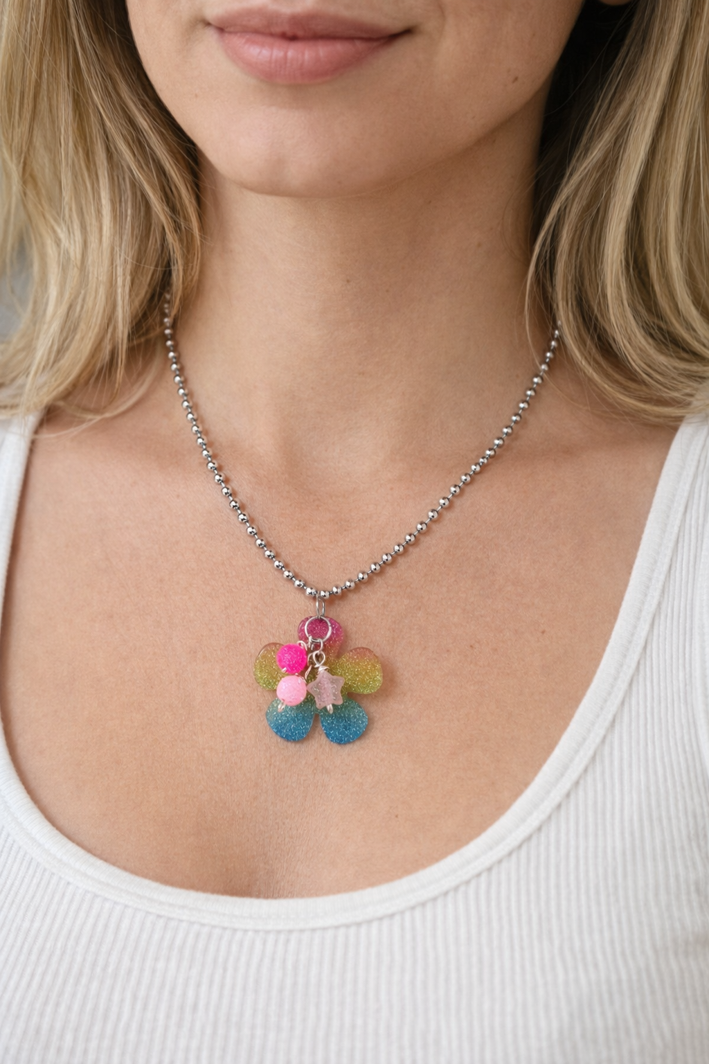 Close-up of a woman wearing a silver beaded necklace with a colorful glitter flower pendant and pink charms, white ribbed top