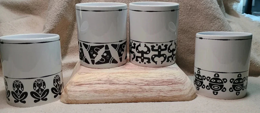 White ceramic mugs with black Taino indigenous patterns on wooden display against beige fabric background